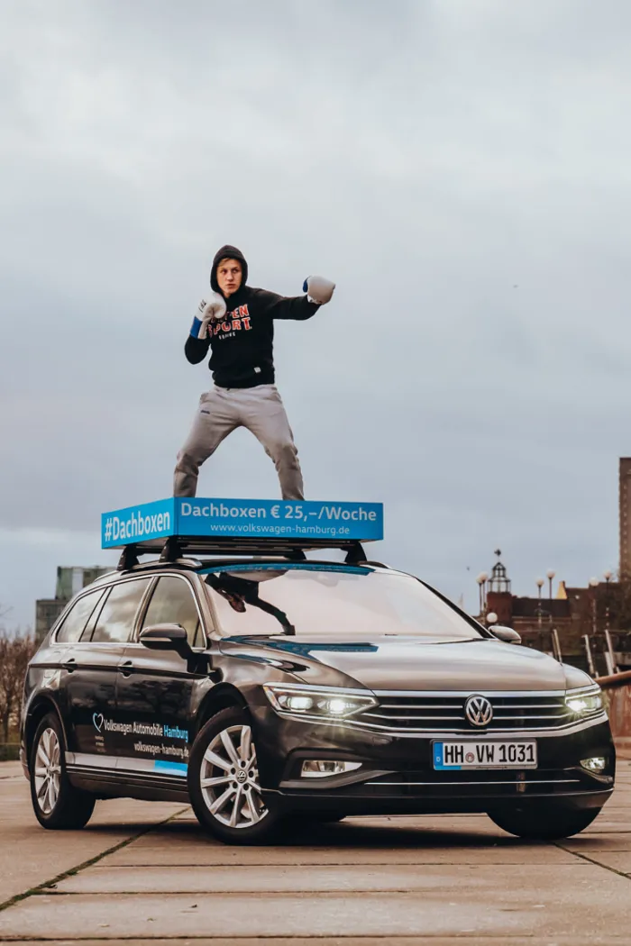 Auf einem VW ist ein Mann als Boxer mit Boxhandschuhen auf einer Fläche abgebildet, die normalerweise für eine Dachbox verwendet wird.
