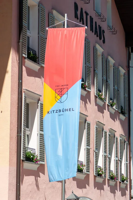 In der Kitzbüheler Innenstadt wurde eine Fahne mit dem neuen Jubiläums-Logo abgebildet. Das Logo in den Farben Rot, Gelb und Blau zieht sich über die gesamte Breite des Banners.
