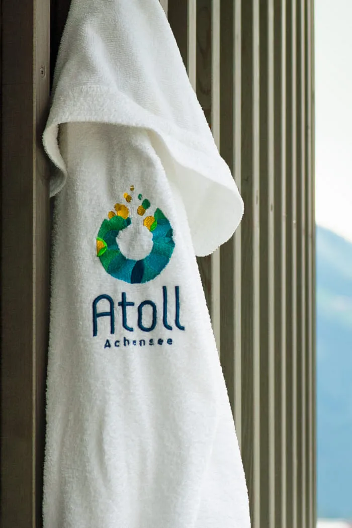 An einem Wandhaken auf einer Holzwand im Außenbereich hängt ein weißer Bademantel, auf dessen Rückseite das Atoll-Logo gestickt ist. Das Logo symbolisiert einen Wassertropfen, darunter steht der Schriftzug 'Atoll Achensee'.