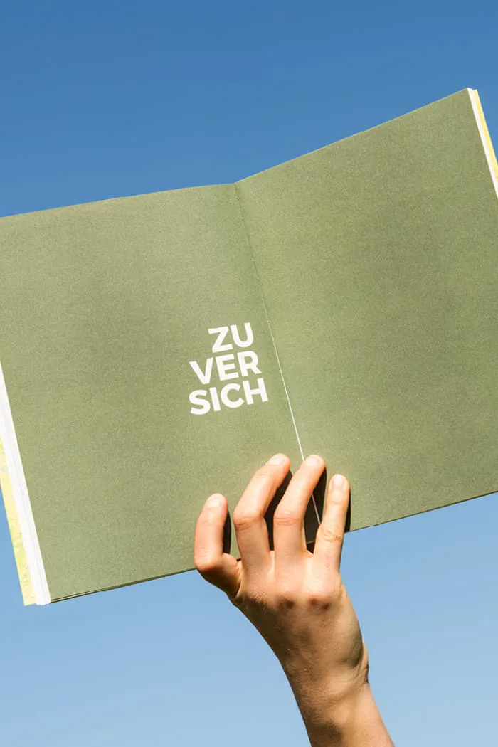 Geöffnete Buchseite mit dem Wort Zuversicht in weißer Typografie auf grünem Hintergrund, gehalten vor strahlend blauem Himmel.