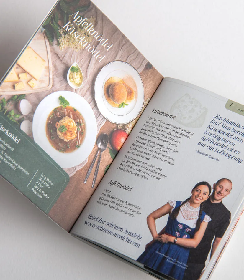Auf weißer Oberfläche liegt das aufgeschlagene Rezeptbuch vom 42. Knödelfest. Die Doppelseite zeigt eine Fotografie der Knödel, rechts daneben ist das Rezept abgedruckt. Zusätzlich ist ein Portraitfoto der Personen zu sehen, die diesen Knödel beim Knödelfest zubereiten.