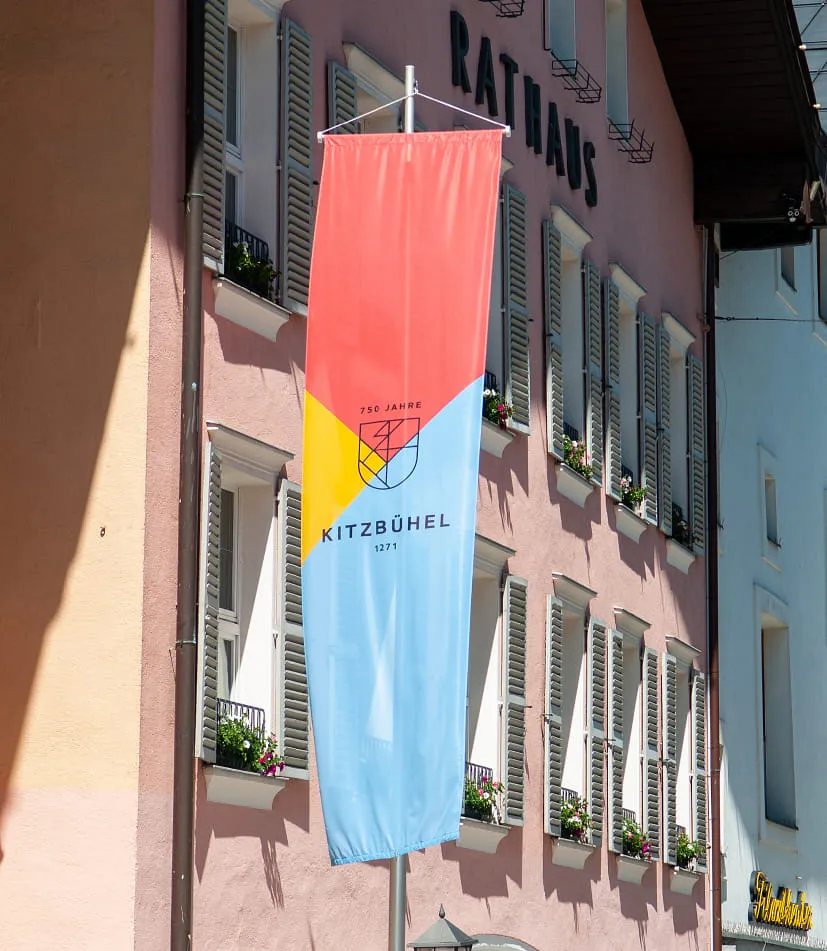 Vor dem Kitzbühler Rathaus mit roter Fassade und vielen Fenstern hängt eine Fahne im Layout des 750 jährigen Jubiläums. In der Mitte befindet sich ein stilisiertes Wappen mit grafischen Linien, darüber steht '750 Jahre', darunter 'KITZBÜHEL 1271'. Der Hintergrund ist in drei farbige Dreiecke unterteilt, oben rot, links gelb, unten blau.
