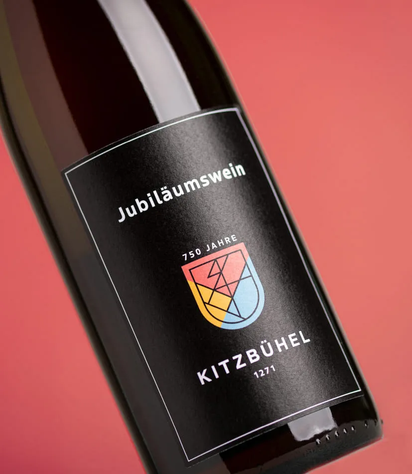 Nahaufnahme einer Weinflasche mit schwarzem Etikett vor rotem Hintergrund. Auf dem Etikett steht 'Jubiläumswein' sowie '750 Jahre Kitzbühel 1271'. In der Mitte befindet sich ein farbiges Wappen mit geometrischem Design in Gelb, Rot und Blau.