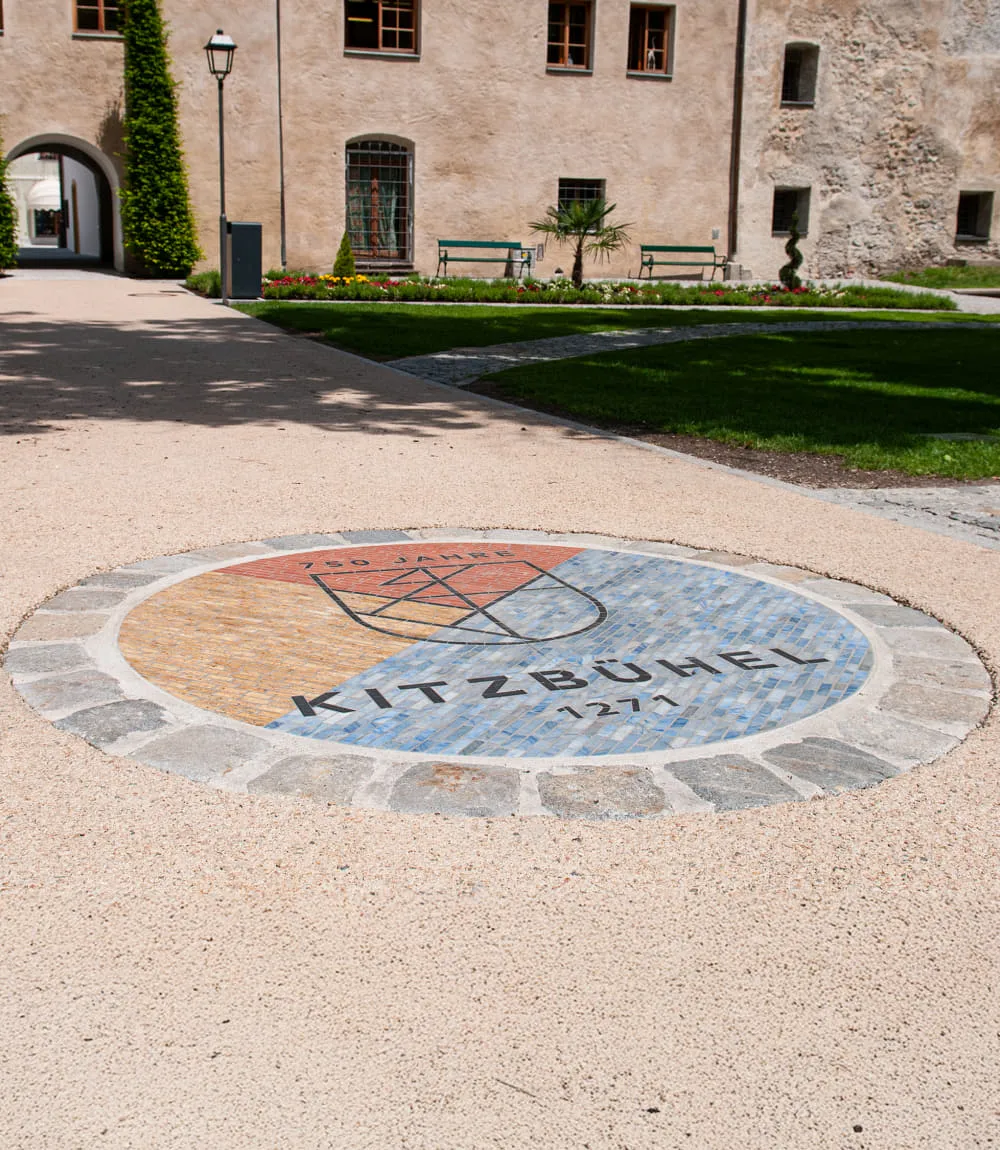 Rundes Bodenmosaik im Innenhof mit dem Jubiläumslogo „750 Jahre Kitzbühel 1271“. Das Mosaik zeigt ein stilisiertes Wappen auf farblich geteiltem Untergrund in Gelb, Rot und Blau. Im Hintergrund sind eine historische Gebäudefassade, grüne Rasenflächen und Parkbänke zu sehen.