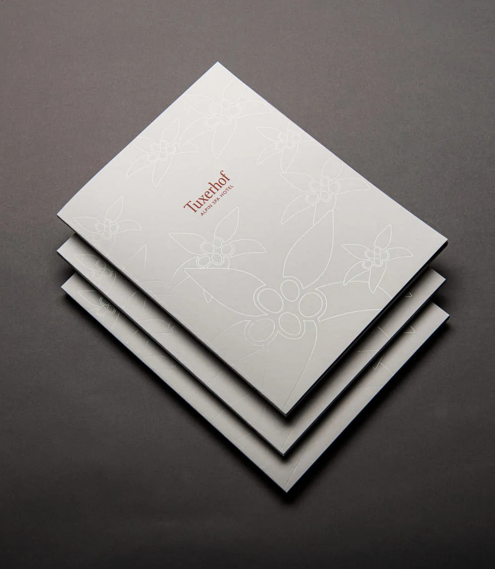 Elegante Broschüren des Tuxerhofs mit weißem Cover und dezenten Blumenprägungen, gestapelt auf dunklem Untergrund. Das Logo Tuxerhof Alpin Spa Hotel ist in Rotbraun zentral auf dem obersten Exemplar platziert.