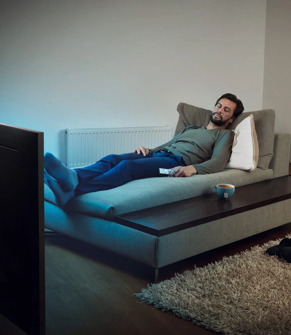Mann entspannt auf grauer Couch im Wohnzimmer vor dem Fernseher, blaues Licht fällt auf die gemütliche Szene mit Tasse auf dem Tisch.