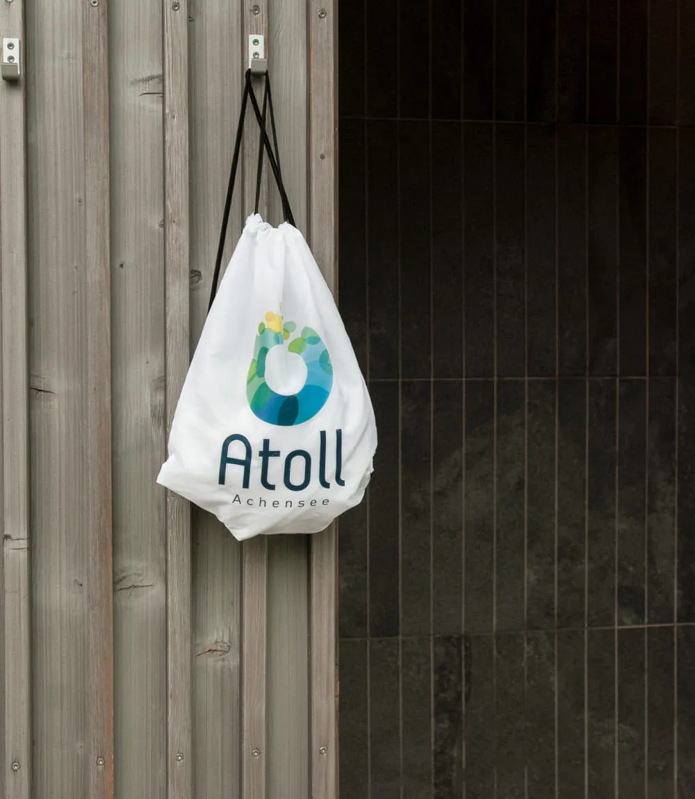 Weißer Sportbeutel mit Atoll Achensee Logo an einer Holzwand hängend.