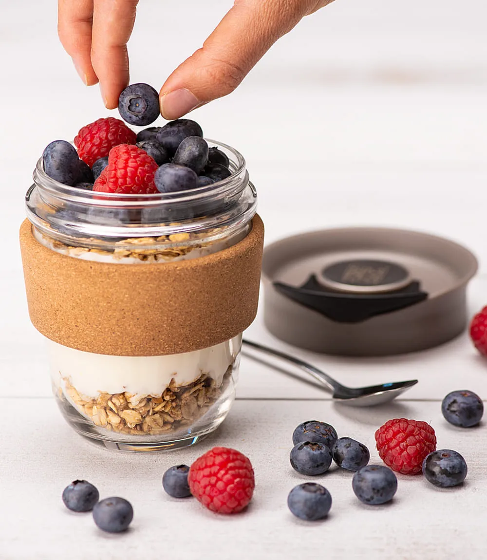 Hand legt Blaubeeren in ein Glas mit Joghurt, Müsli und Korkmanschette.