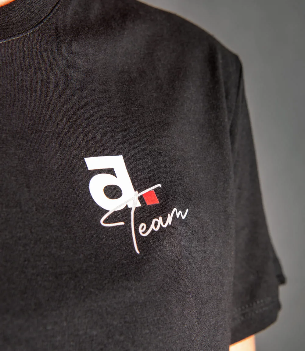 Vordere rechte Seite eines schwarzen T-Shirts vor grauem Hintergrund mit Schriftzug Team in weiß und kleinem weißen a mit viereckigem roten Punkt.