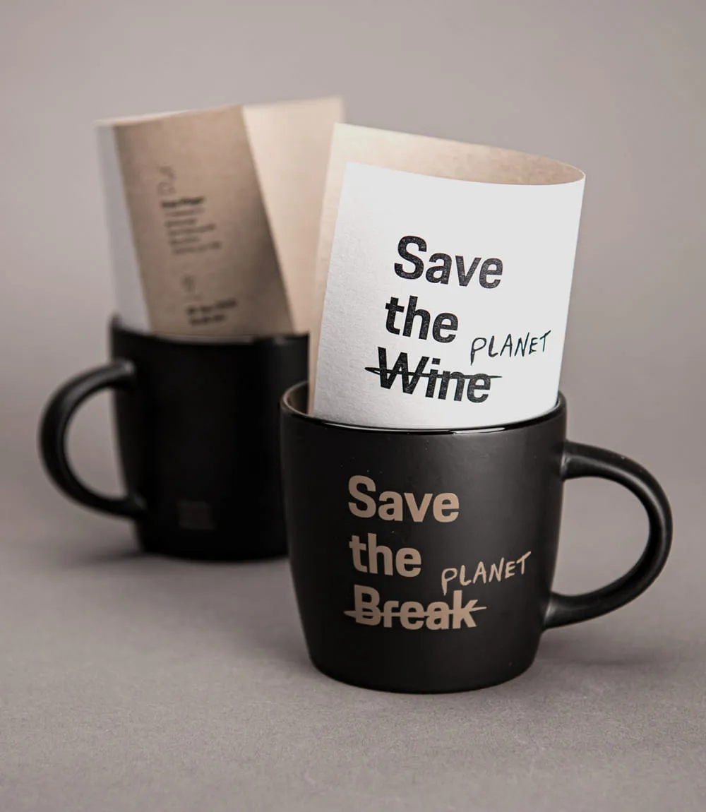 Zwei schwarze Tassen mit goldener Aufschrift stehen auf einem grauen Hintergrund. In der vorderen Tasse steckt ein gefaltetes Blatt mit dem Schriftzug Save the Wine – Wine durchgestrichen und ersetzt durch Planet, auf der Tasse selbst steht Save the Break – wiederum ersetzt durch Planet.