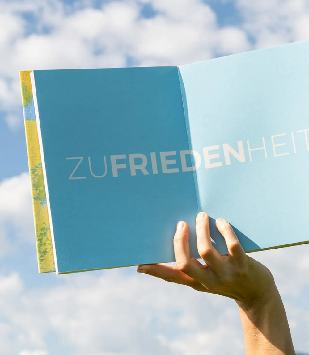 In der Mitte geöffnetes Buch wird von einer Hand vor wolkenbedecktem Himmel gehalten. Aufgeschlagene Seite zeigt in weißen Großbuchstaben auf hellblauem Hintergrund das Wort Zufriedenheit wobei Frieden fettgedruckt ist.