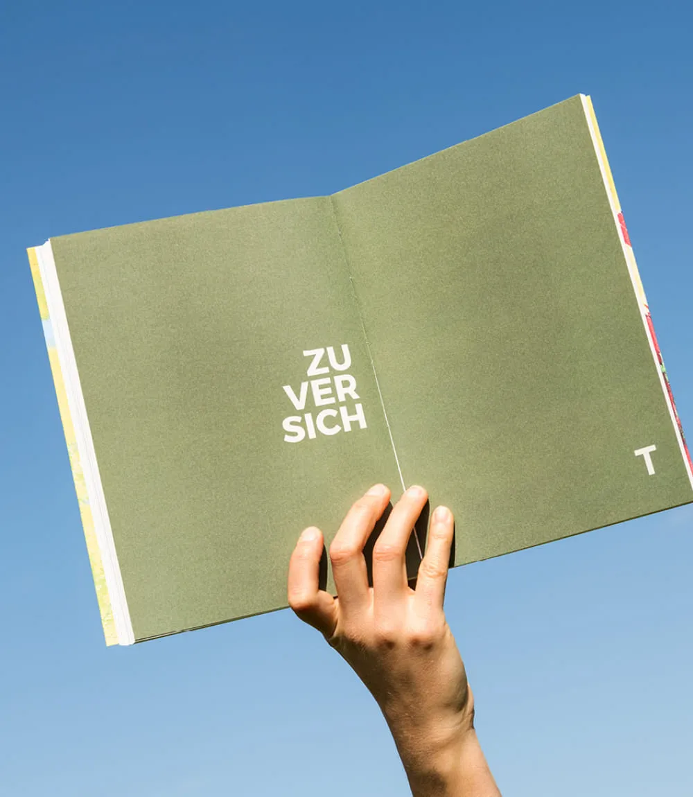 Geöffnete Buchseite mit dem Wort Zuversicht in weißer Typografie auf grünem Hintergrund, gehalten vor strahlend blauem Himmel.