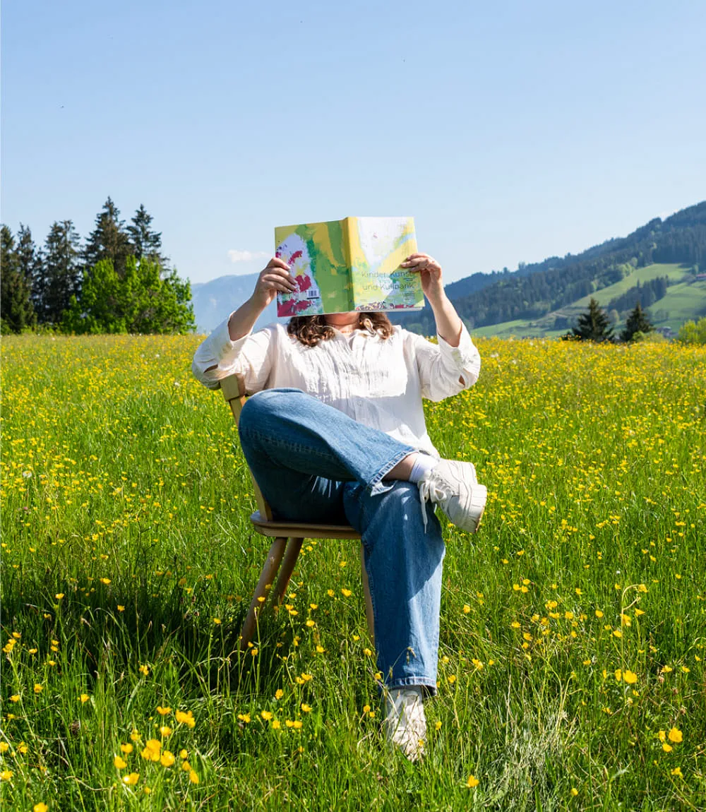 Person sitzt entspannt auf einem Holzstuhl inmitten einer blühenden Sommerwiese voller gelber Blumen. Sie trägt Jeans, weiße Sneakers und eine helle Bluse. Das Gesicht ist durch das aufgeschlagene, farbenfrohe Rezeptbuch Kinder, Kunst und Kulinarik verdeckt. Im Hintergrund sind grüne Hügel, Wälder und ein klarer blauer Himmel mit Alpenpanorama zu sehen.