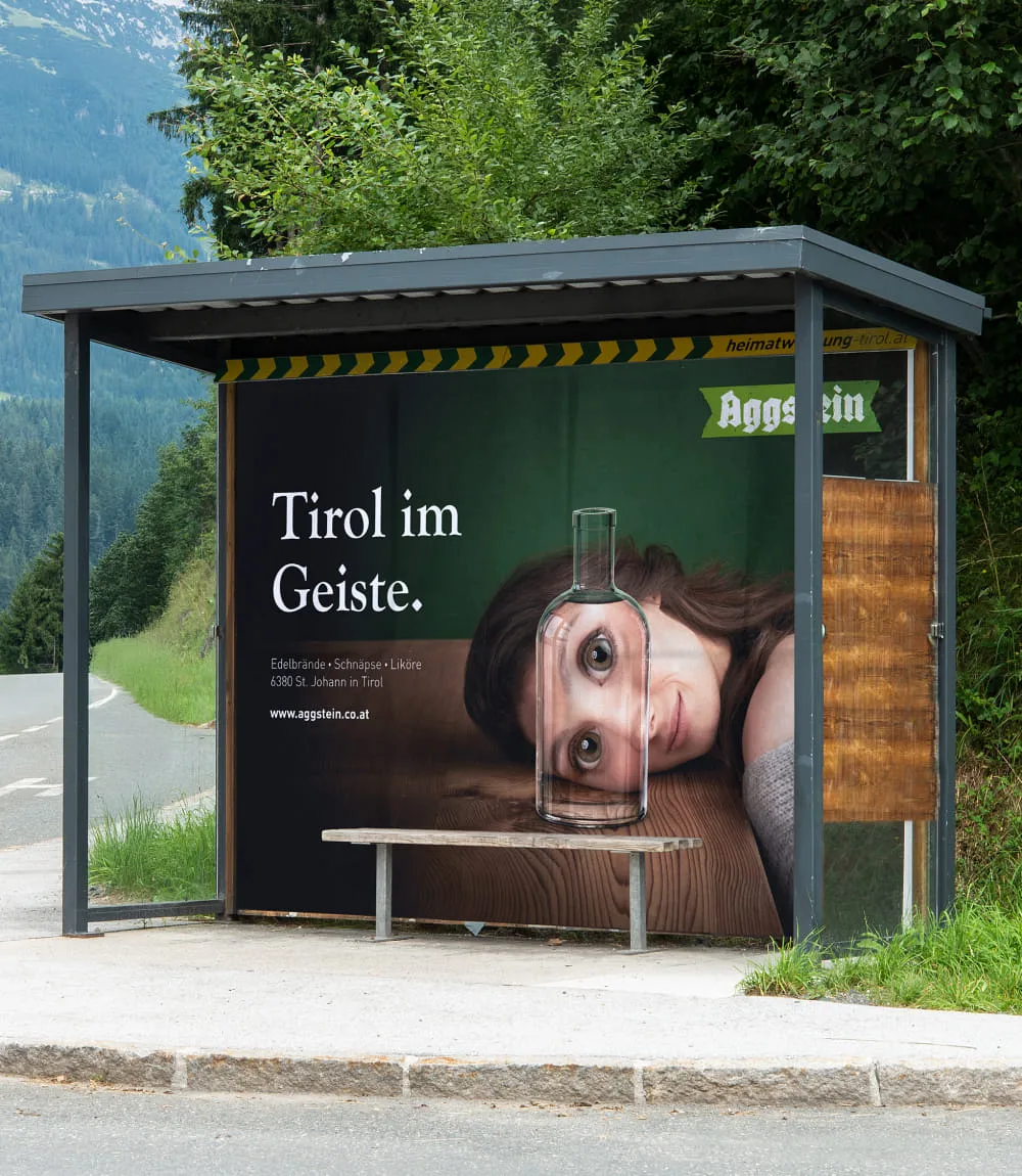 In einem Bushaltestellenhäuschen ist ein großformatiges Werbeplakat von Aggstein zu sehen. Darauf eine junge Frau mit braunen Haaren, die ihren Kopf auf einem Holztisch liegend, durch eine Glasflasche blickt in der ihre Augen stark vergrößert erscheinen. Links eine Schriftblock in weiß betitelt mit Tirol im Geiste.
